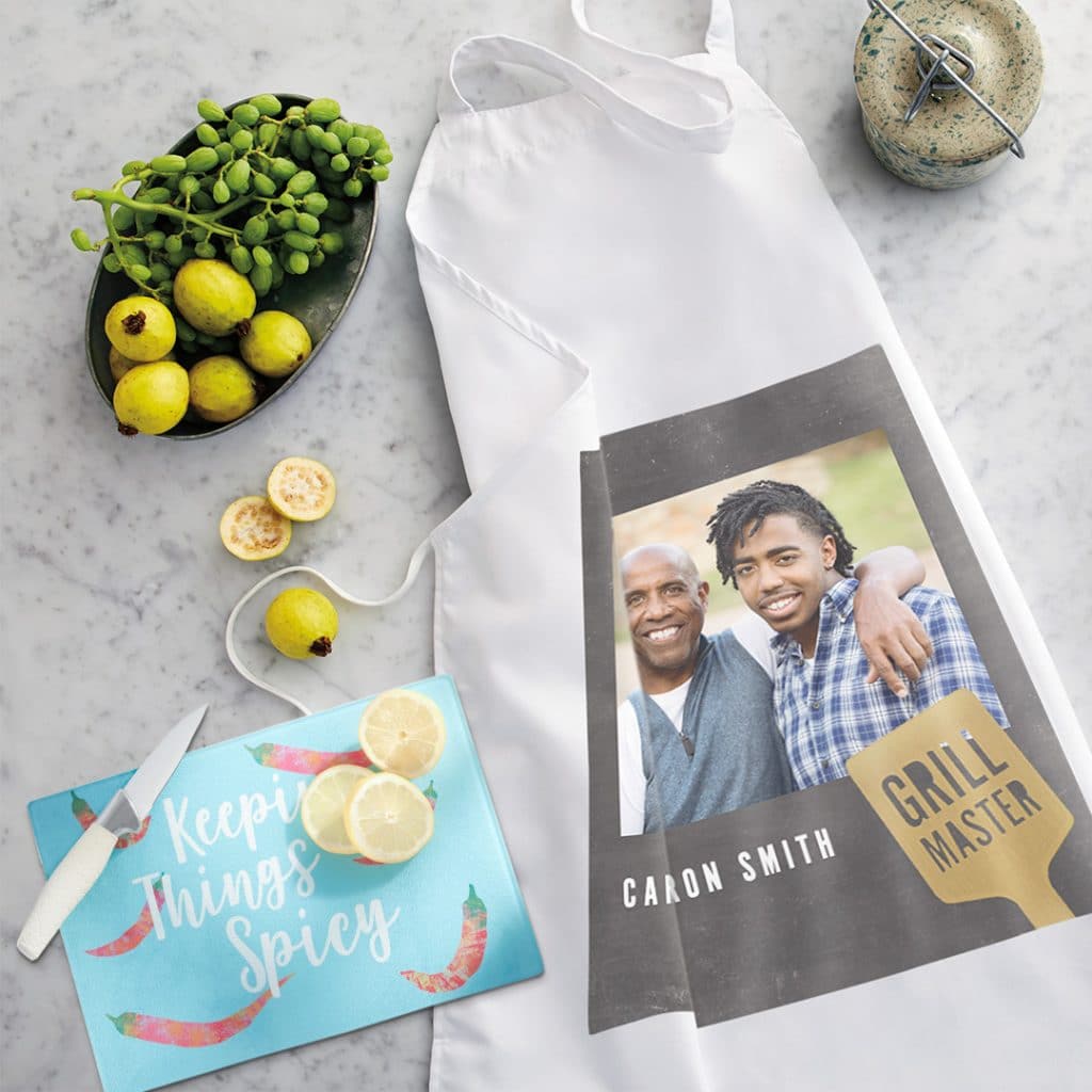 Custom apron featuring dad and son photo laying on a countertop next to a glass cutting board that reads "Keeping Things Spicy"