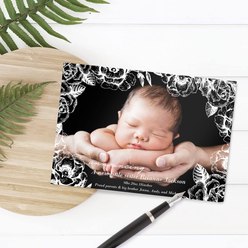 Baby Roses Overlay birth announcement card design laying over plant leaves on a table, alongside a black pen