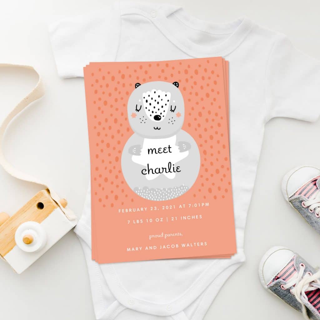 Bear Snuggle baby announcement card design laying over a newborn babysuit, alongside little shoes and a toy