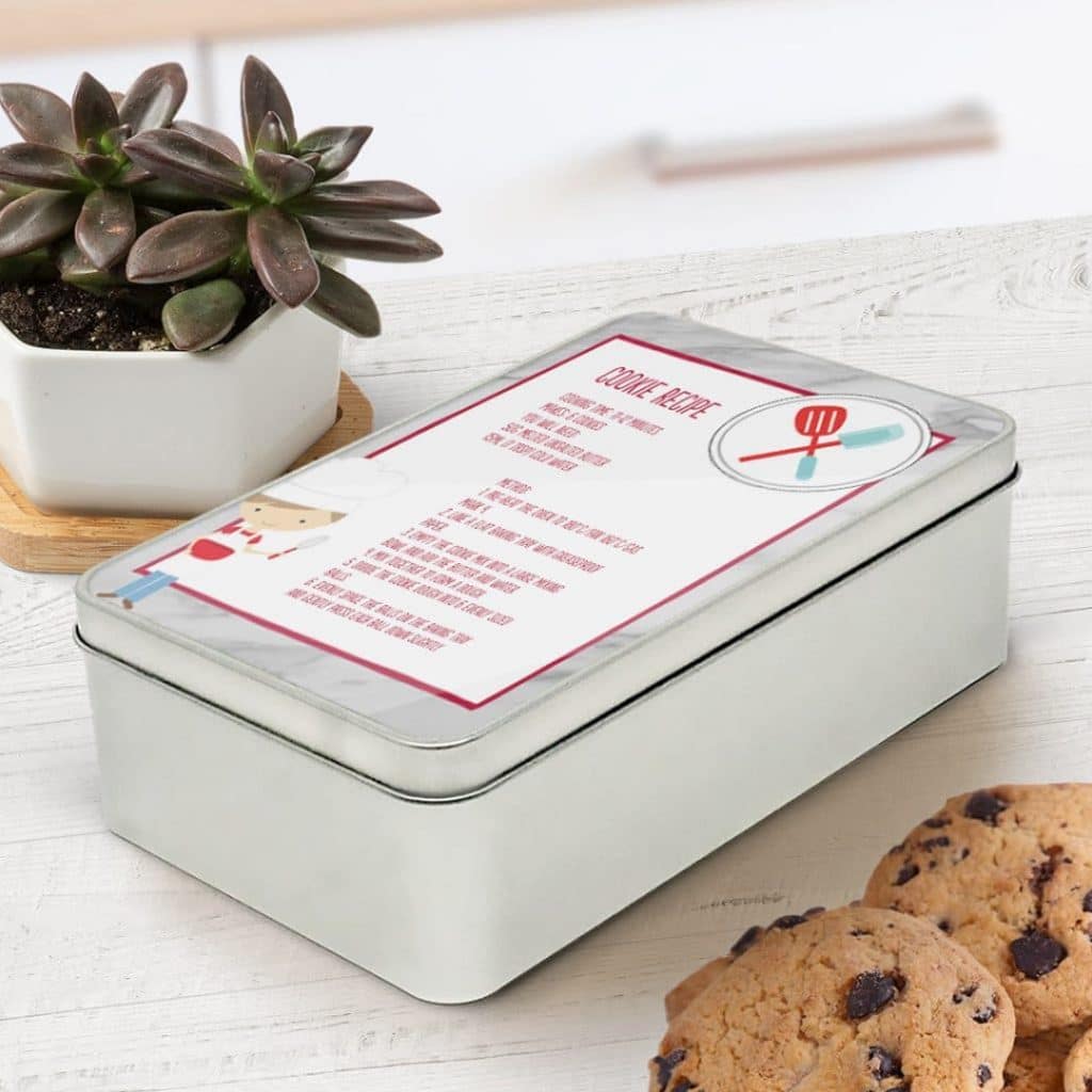 A rectangle tin with a cookie recipe on the lid, next to some chocolate chip cookies