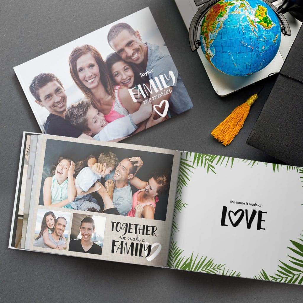 A closed and an open photo book with family images shown on a dark grey surface next to a laptop and a globe