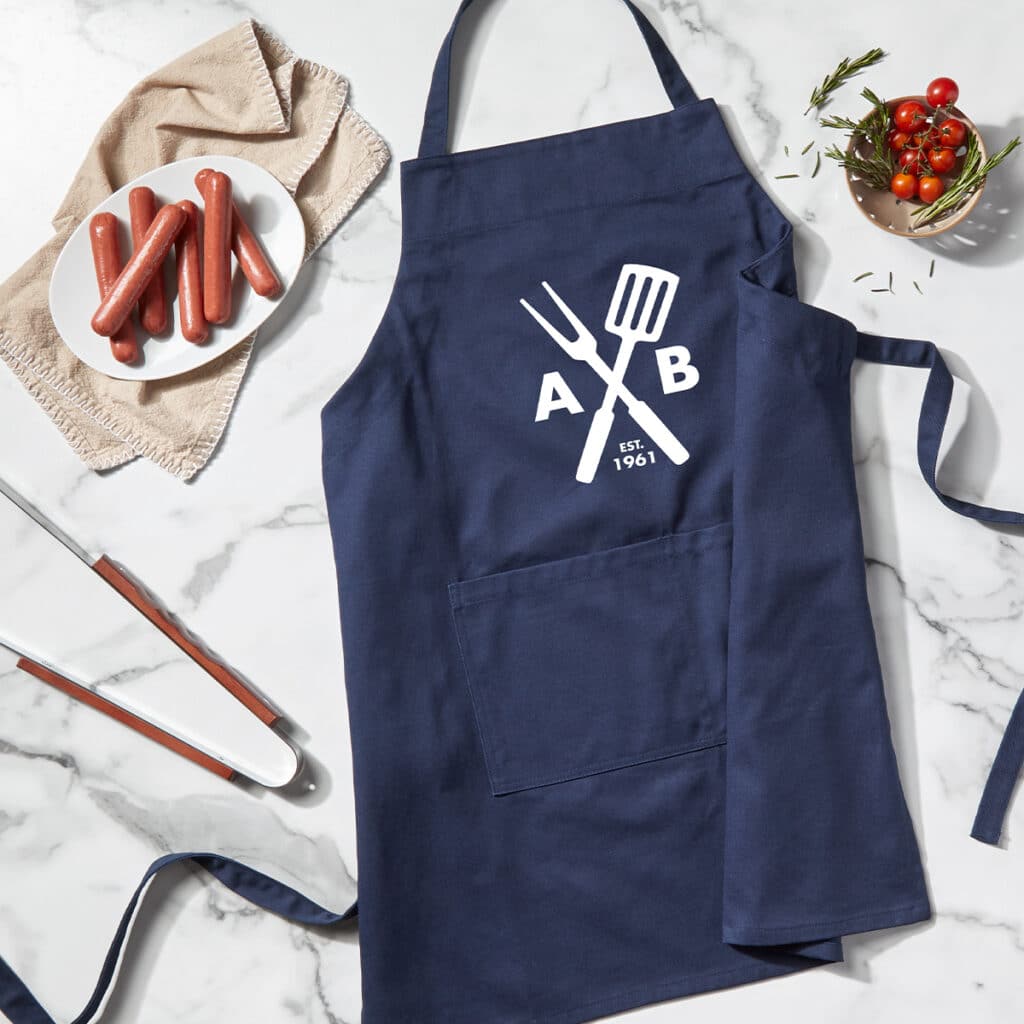 Flat lay image of a navy apron with a monogram grill design. The apron is laying next to grill tools, a plate of hot dogs and a bowl of tomatoes